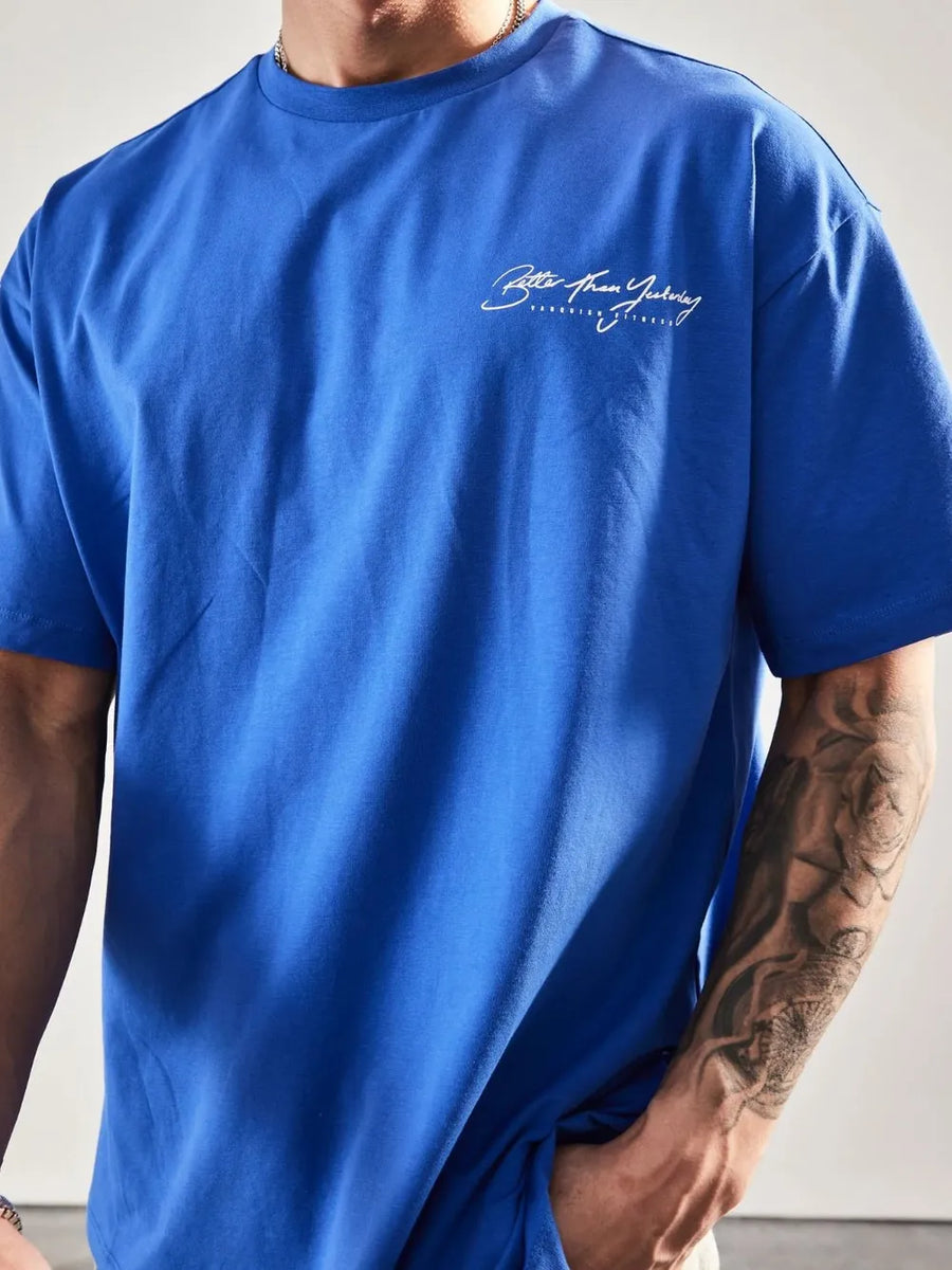 Blue oversized graphic tee with white script print on chest and tattoo on arm.