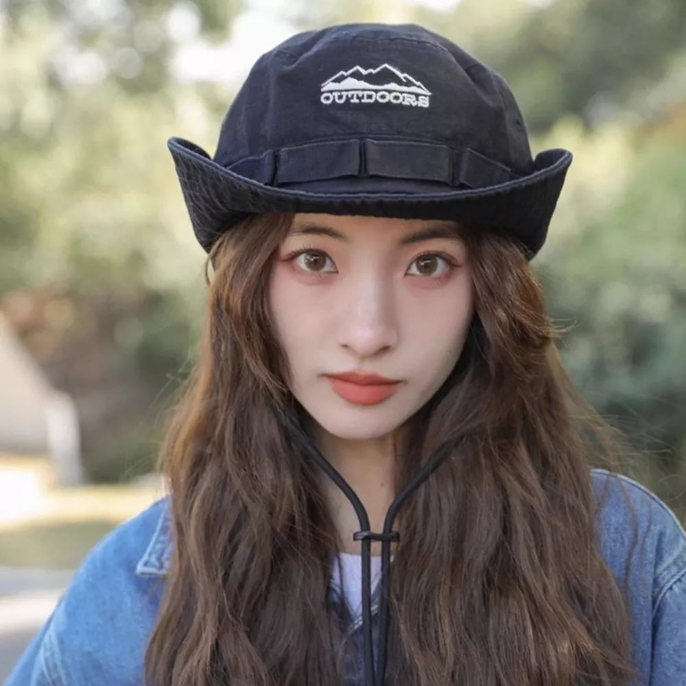 Black bucket hat with embroidered mountain logo and chin strap, worn by a woman in a denim jacket.