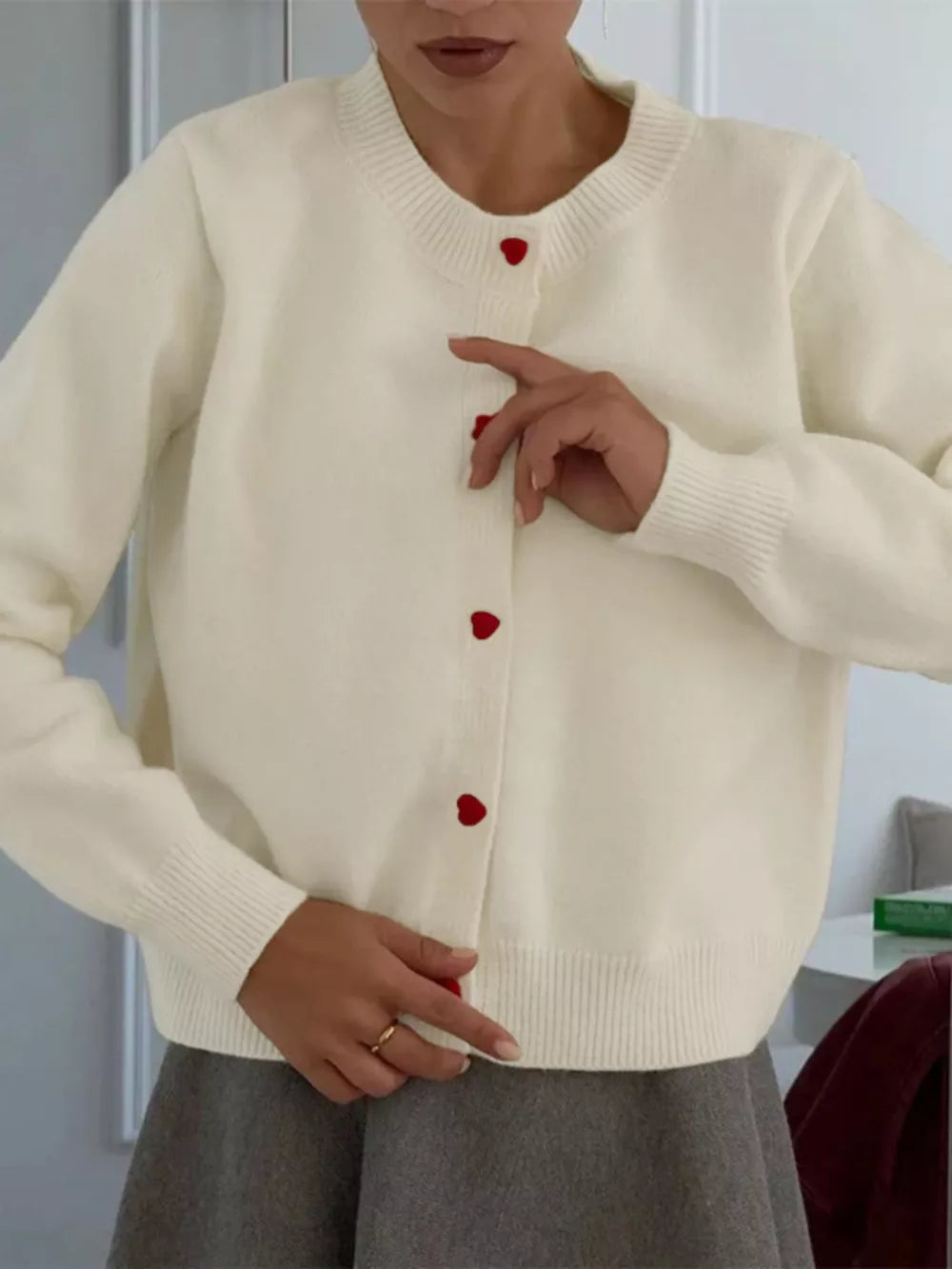 Cream knit cardigan with red heart buttons, crew neck, long sleeves, and ribbed trim.