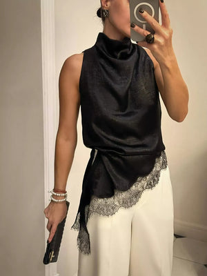 Black asymmetrical cami top with lace trim and cowl neck, styled with white pants.