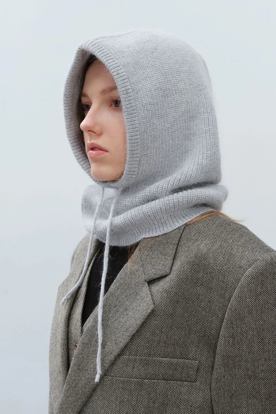 Light gray knit balaclava with hood and drawstring, worn over a blazer.