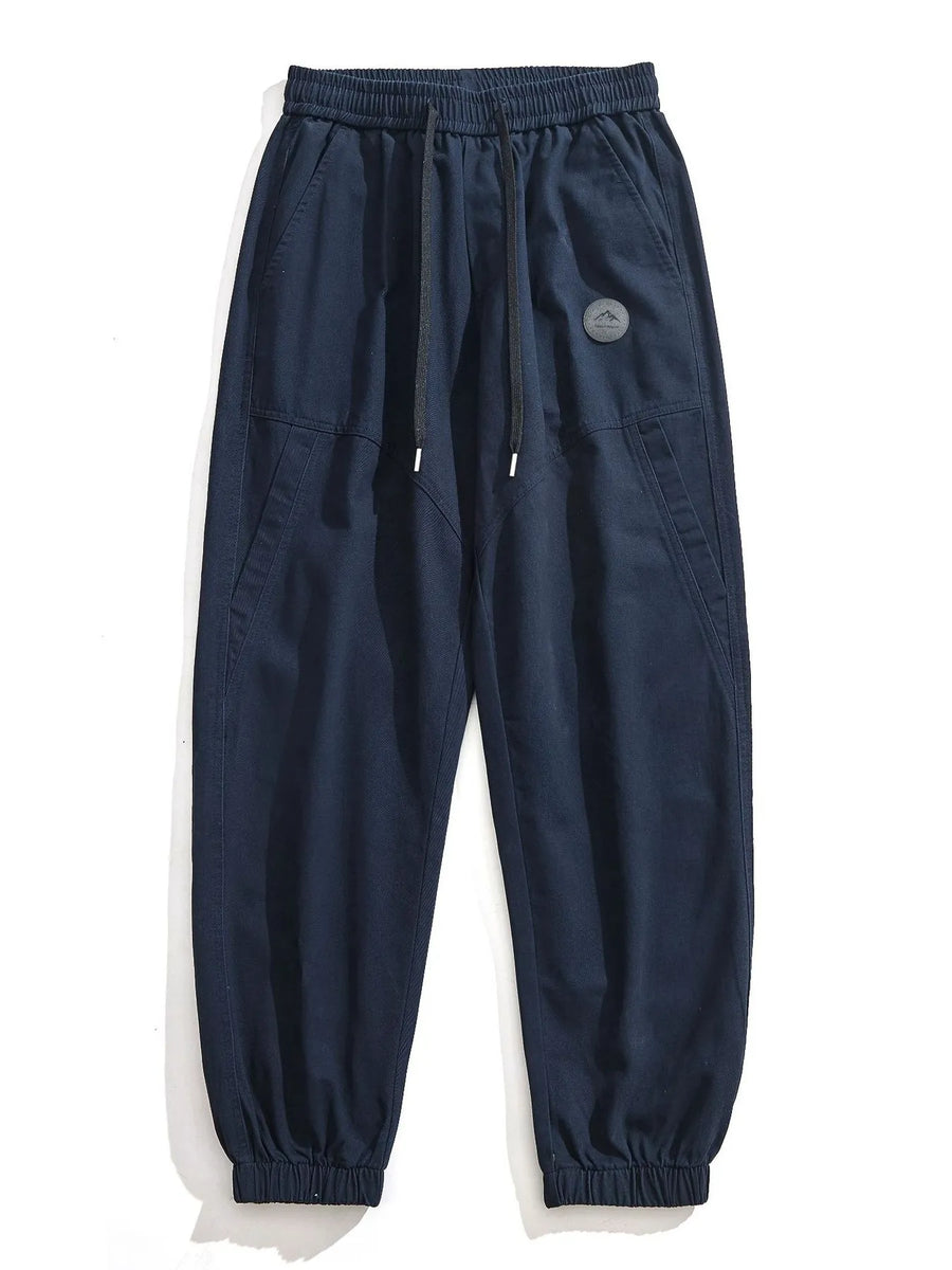 Navy blue relaxed fit joggers with drawstring and elastic cuffs, featuring a circular mountain logo patch.