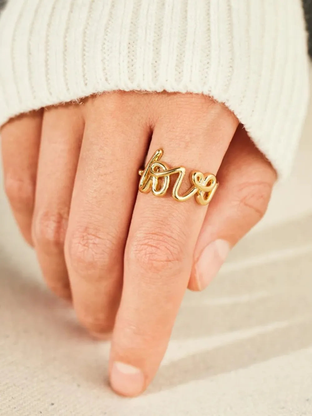 Gold script love ring with a heart detail on a hand wearing a white sweater.