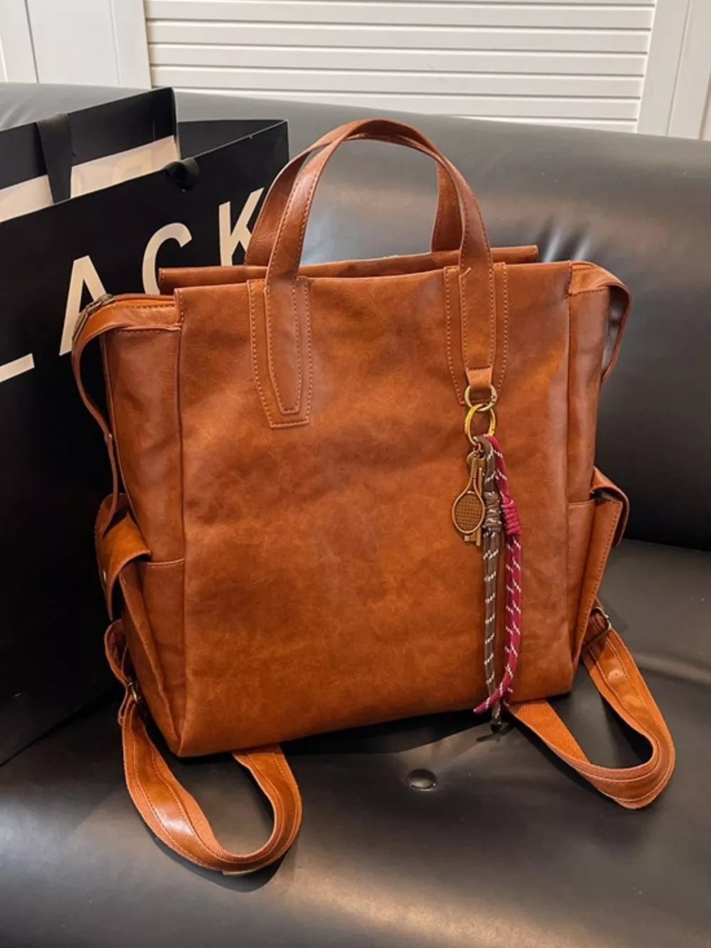 Terracotta leather convertible backpack with top handles and side pockets, featuring a tennis racket charm.
