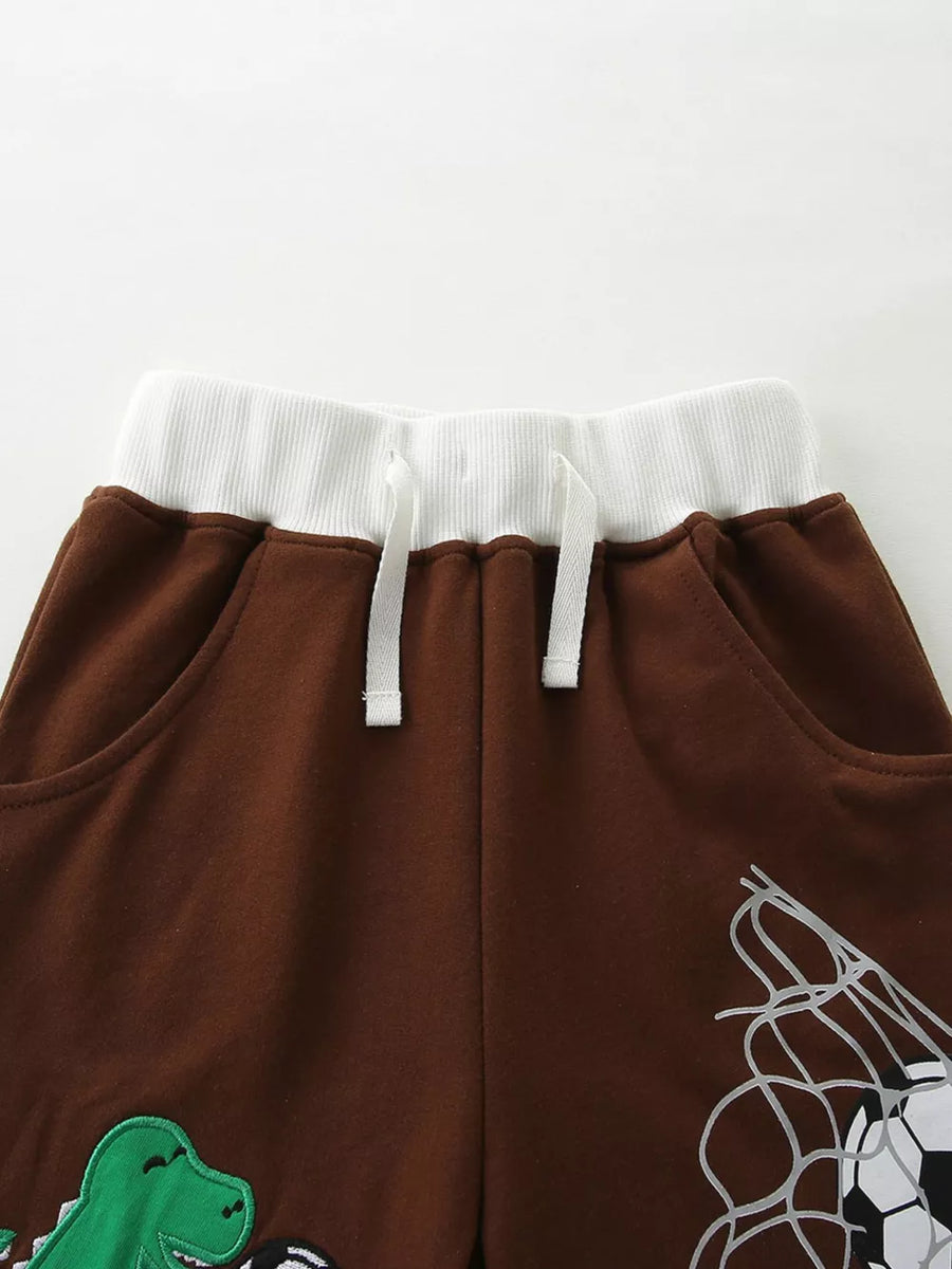 Brown cotton baby shorts with white waistband, drawstring, and graphic soccer ball and dinosaur details.