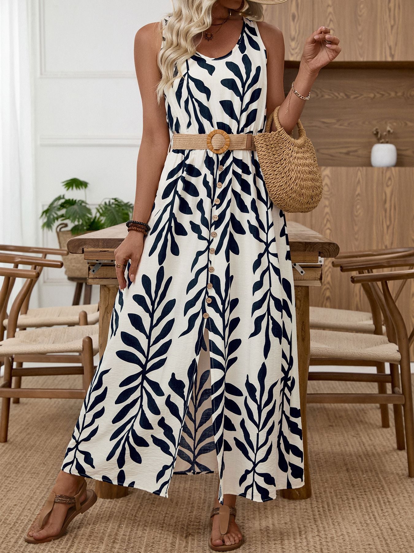 White sleeveless maxi dress with navy tropical leaf print, belted waist, and decorative buttons.