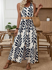 White sleeveless maxi dress with navy tropical leaf print, belted waist, and decorative buttons.