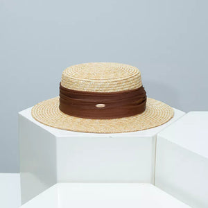 Refined straw boater hat in beige with a brown ribbon detail and small metal emblem.