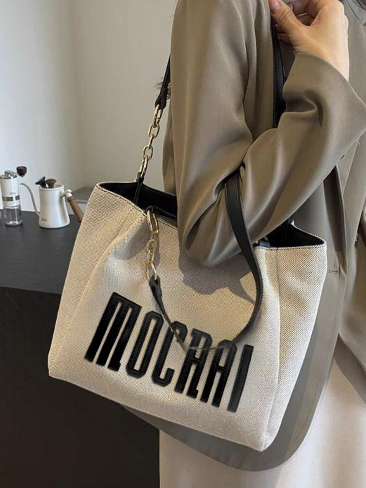 Textured canvas shoulder bag in beige with black chain accents and bold lettering.