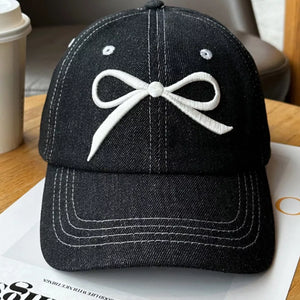 Black denim baseball cap with white embroidered bow accent and contrast stitching.