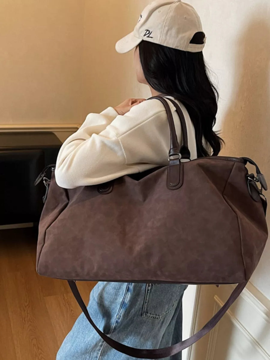 Chocolate duffel bag with adjustable strap and leather handles, worn by a woman.
