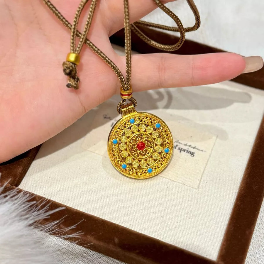 Gold-plated round pendant necklace with red and blue stones on a braided cord.