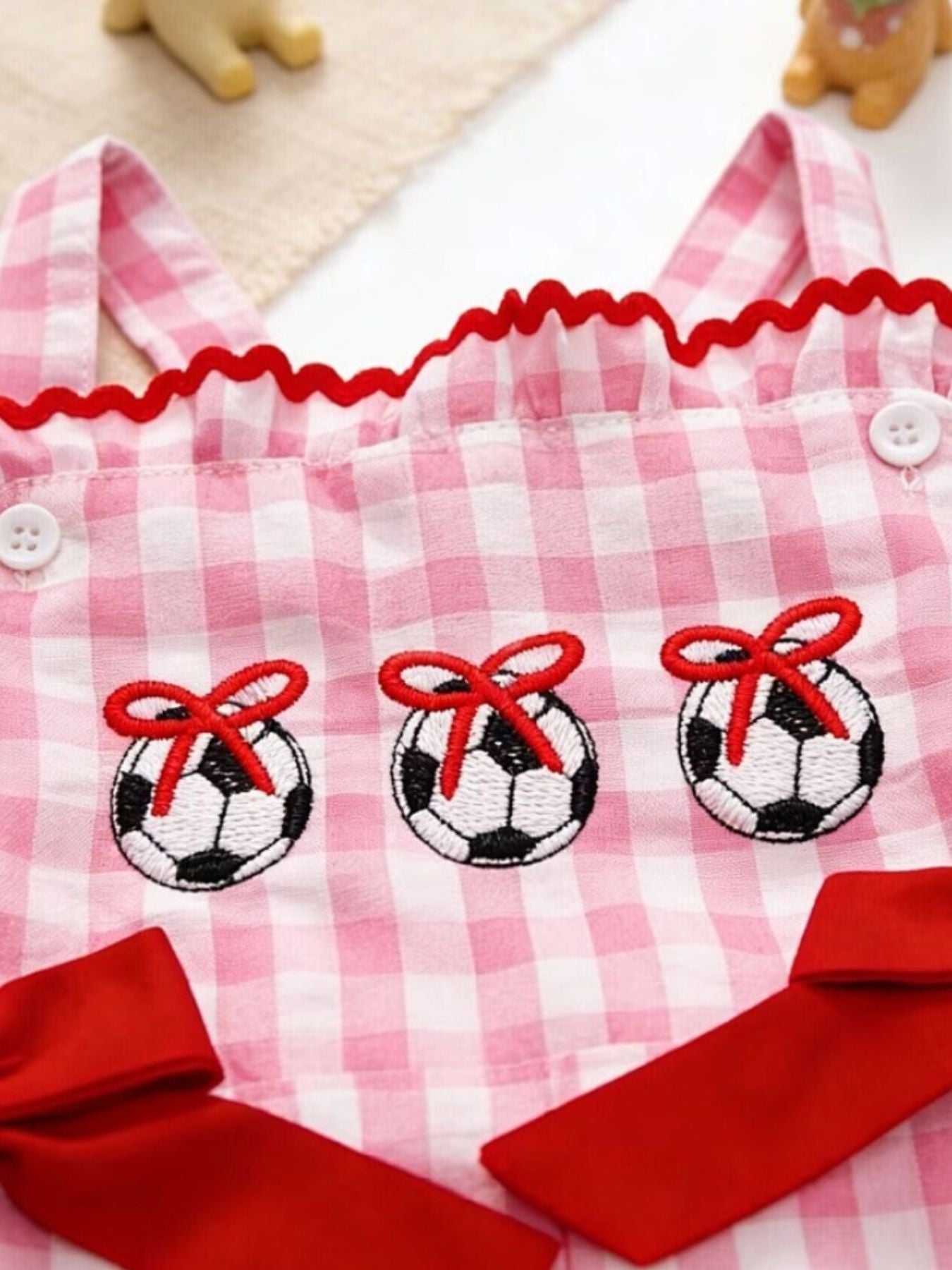 Gingham soccer ball embroidered jumpsuit in blush pink cotton with red trim and bows.