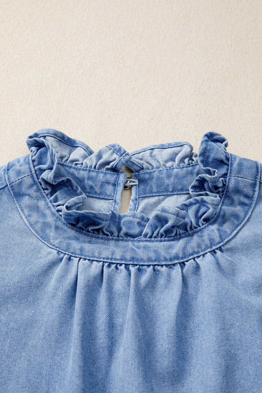 Light wash denim blouse with ruffled collar and button closure at back neck.