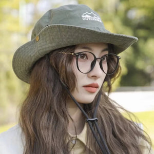 Olive green bucket hat with embroidered mountain logo and chin strap, worn by a woman with glasses.