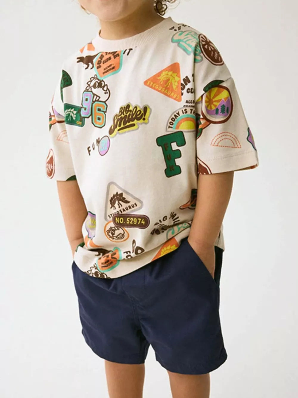 Beige cotton t-shirt with allover dinosaur and adventure patches for toddlers.