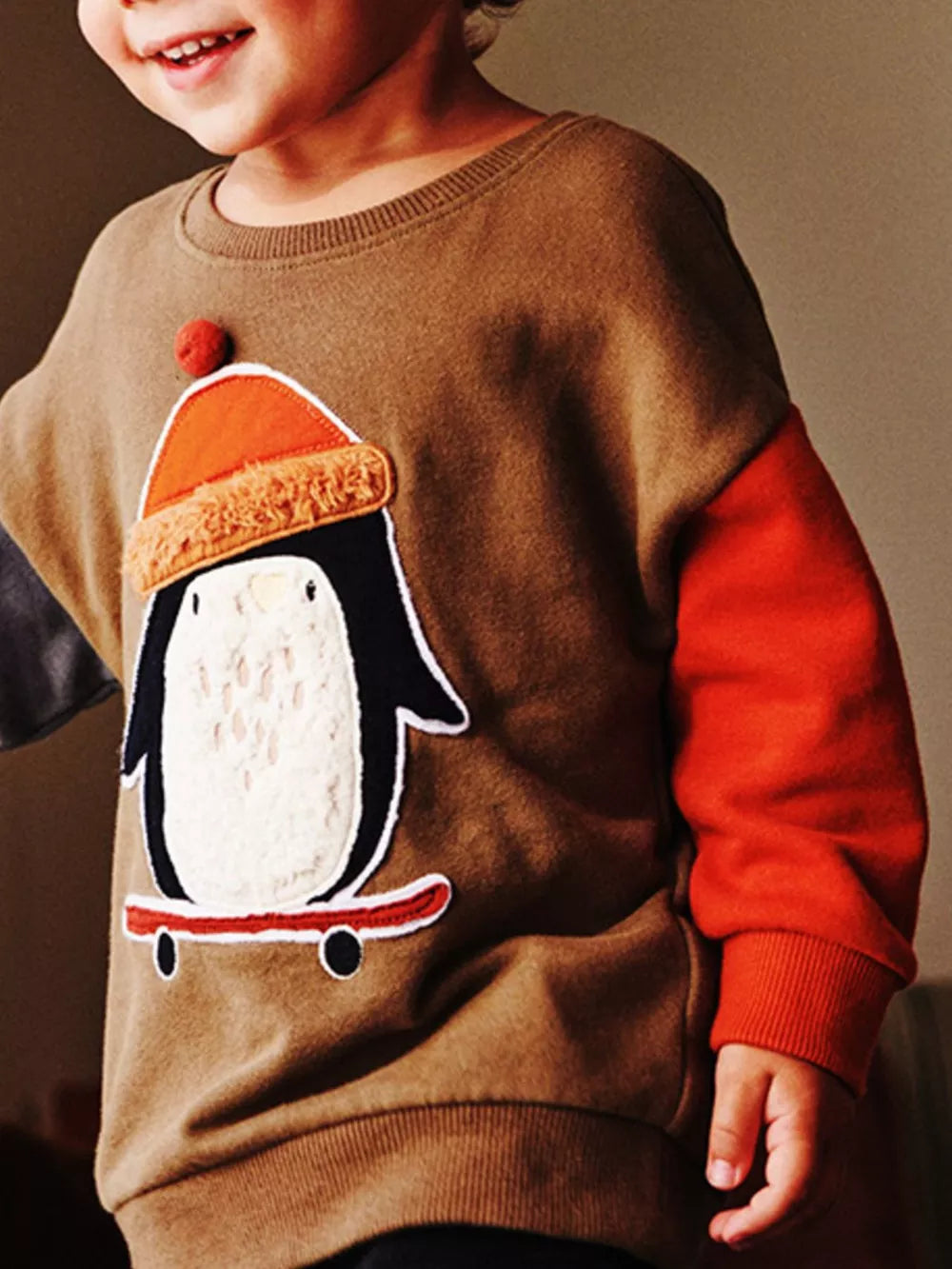 Toddler brown cotton sweatshirt with penguin appliqué on skateboard, orange sleeves.