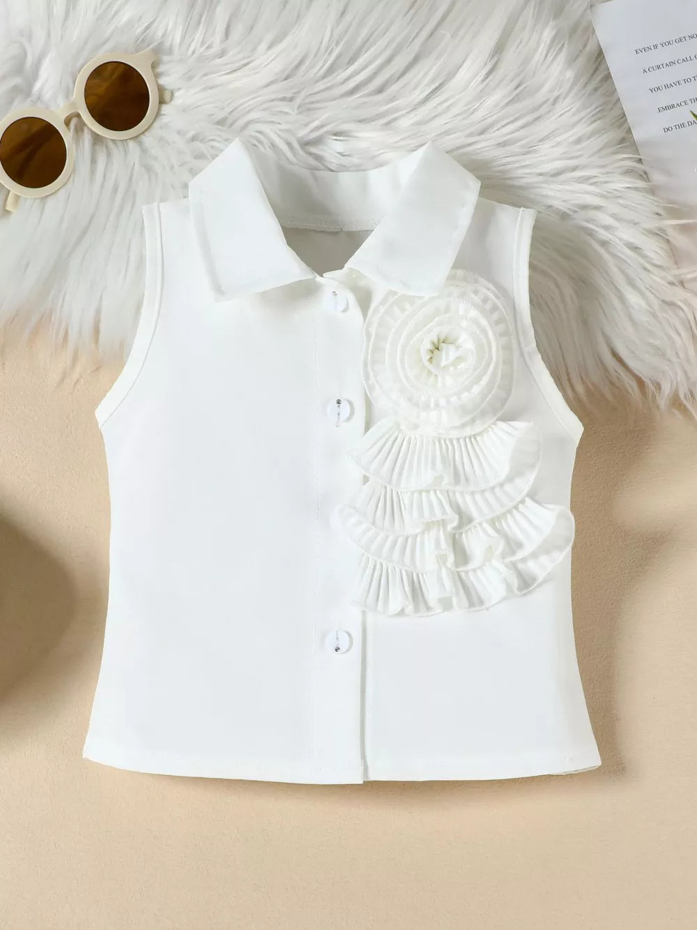 White sleeveless collared blouse with ruffled flower applique and decorative buttons.