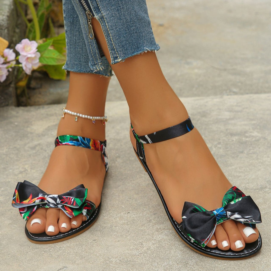 Floral bowknot flat sandals in black with colorful print and ankle strap.