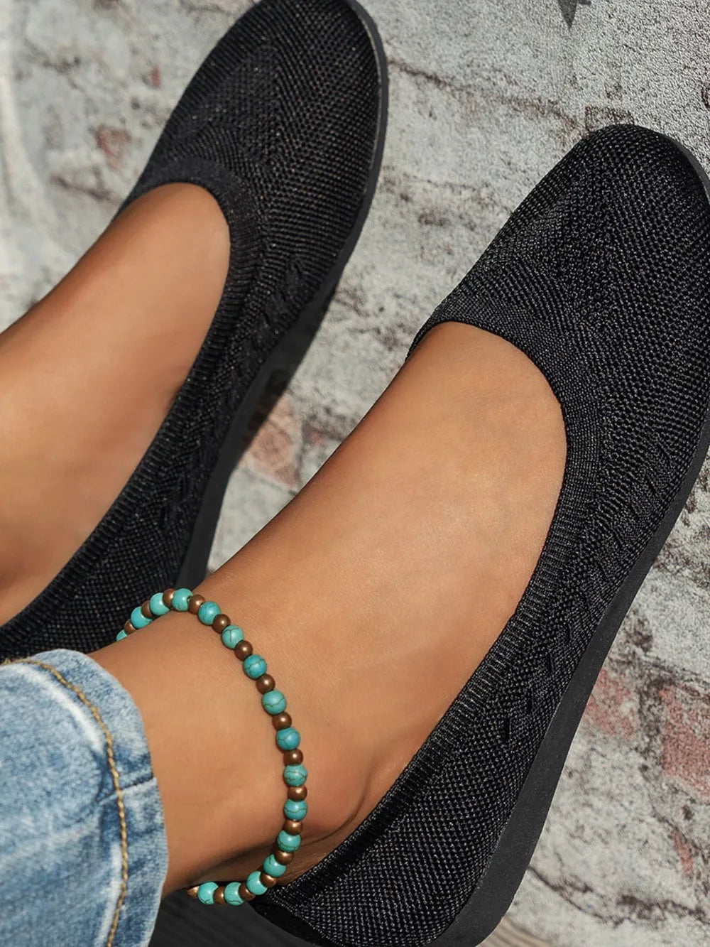 Black textured knit ballet flats with a braided detail, worn with jeans and an anklet.