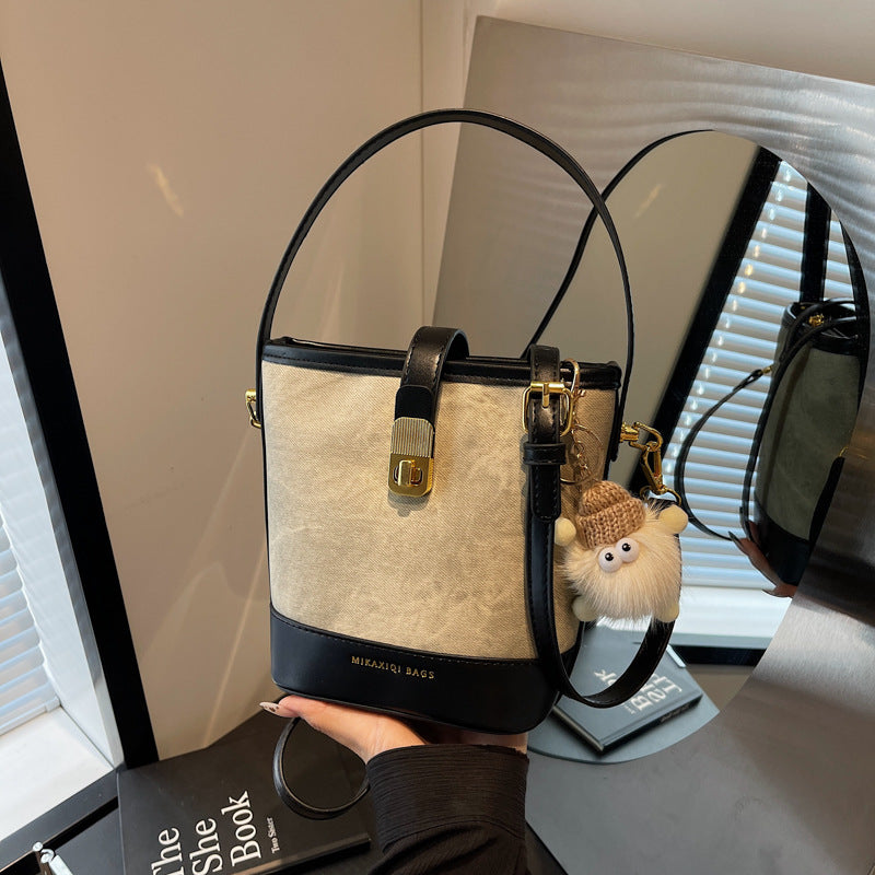 Bucket crossbody bag in beige canvas with black trim and a fuzzy charm.