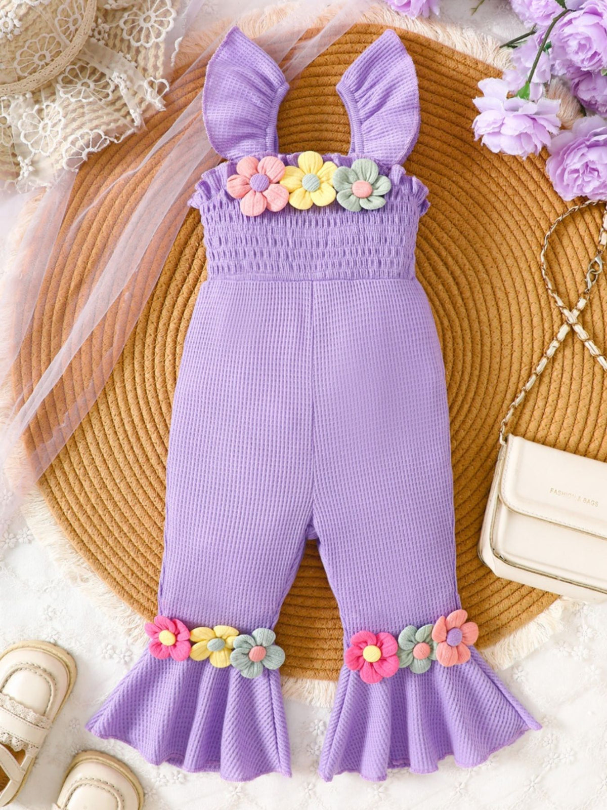 Purple baby jumpsuit with smocked bodice, ruffle straps, and floral applique on neckline and flared legs.
