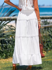 White tiered maxi skirt with smocked waist and ruffled hem, styled with a woven bag.
