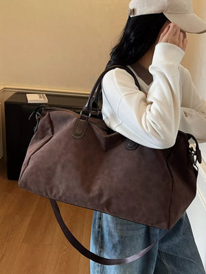 Chocolate duffel bag with adjustable strap and leather handles, worn by a woman.