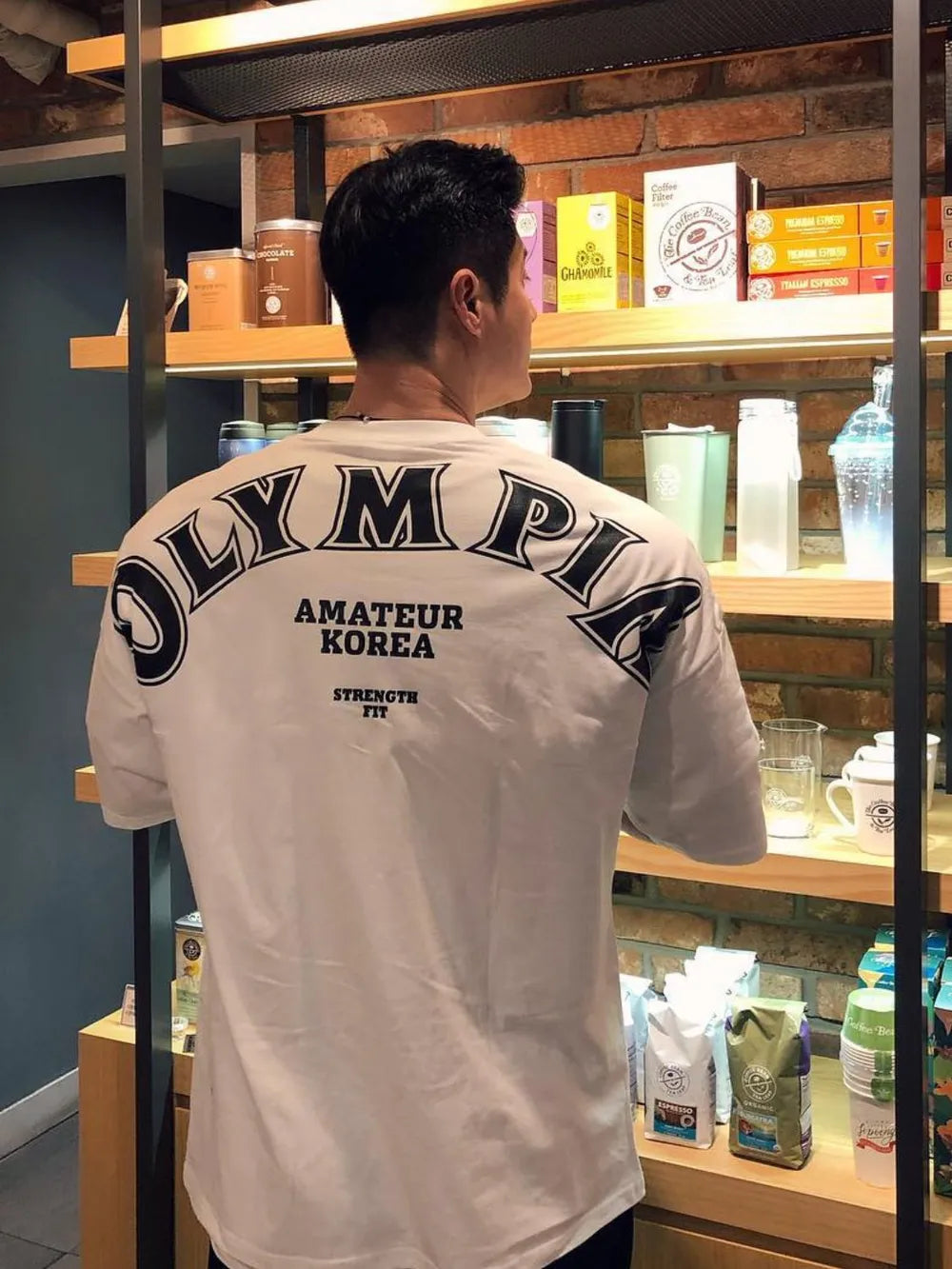 White oversized t-shirt with black "OLYMPIA AMATEUR KOREA STRENGTH FIT" graphic print on back