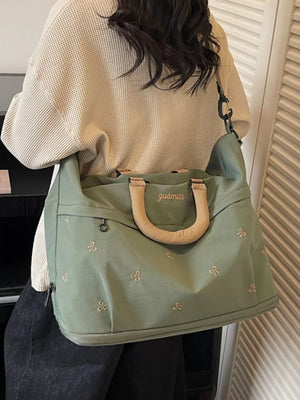 Green duffel bag with embroidered bows and tan handles, worn by a person in a cream waffle knit sweater.