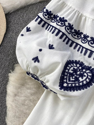White tie-neck blouse with balloon sleeves featuring intricate navy embroidery, including hearts and floral motifs.