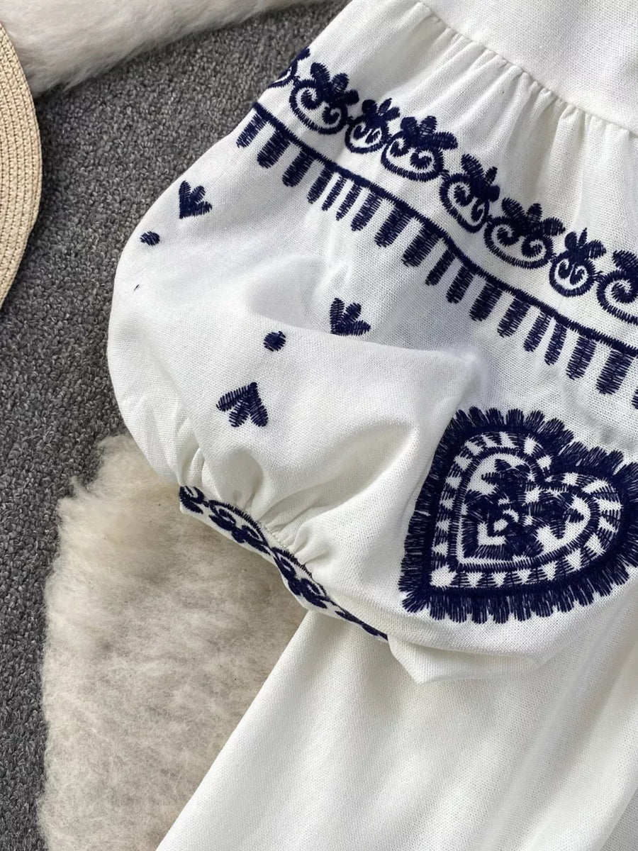 White tie-neck blouse with balloon sleeves featuring intricate navy embroidery, including hearts and floral motifs.