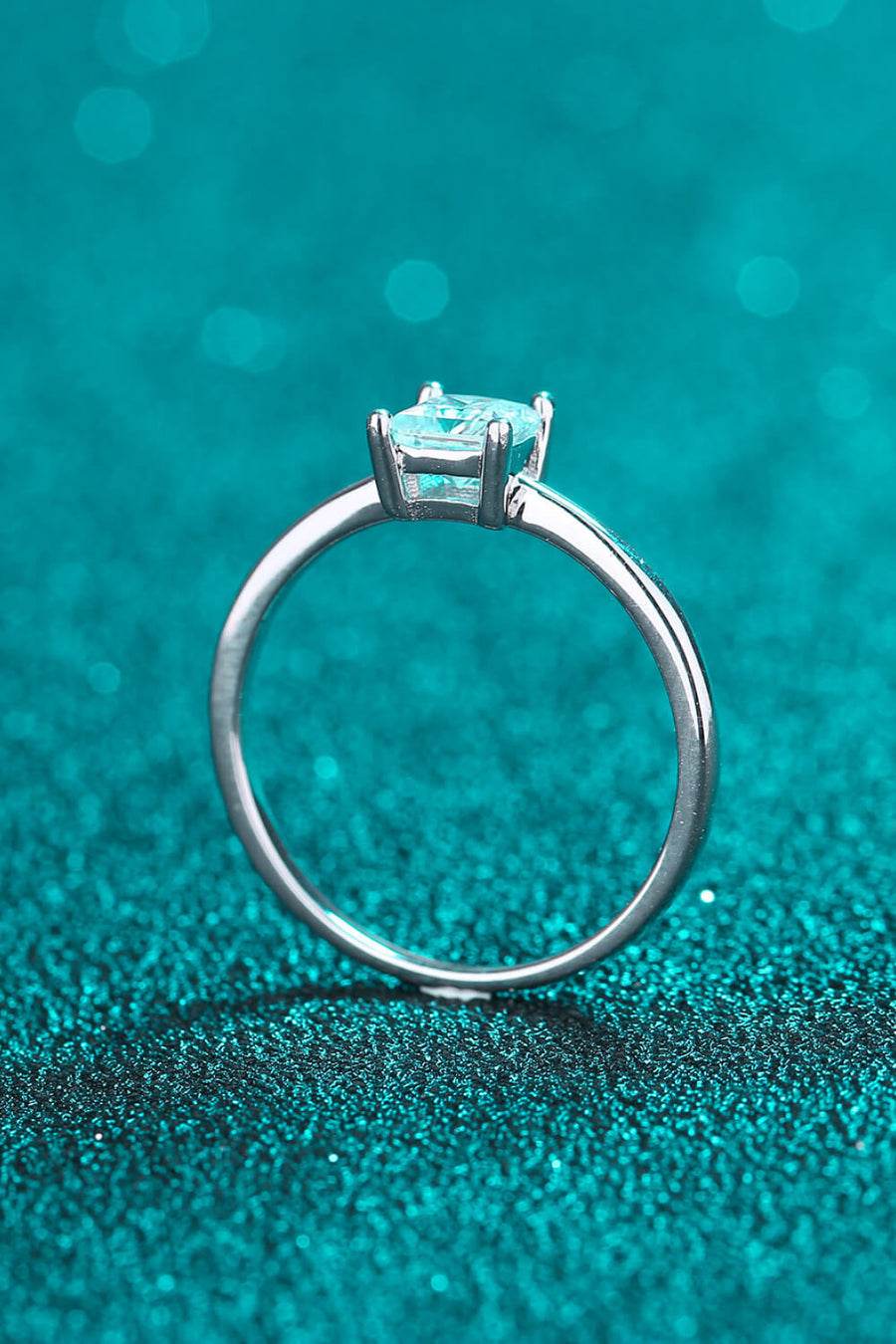Silver ring with a square cut moissanite stone on a teal glitter background.