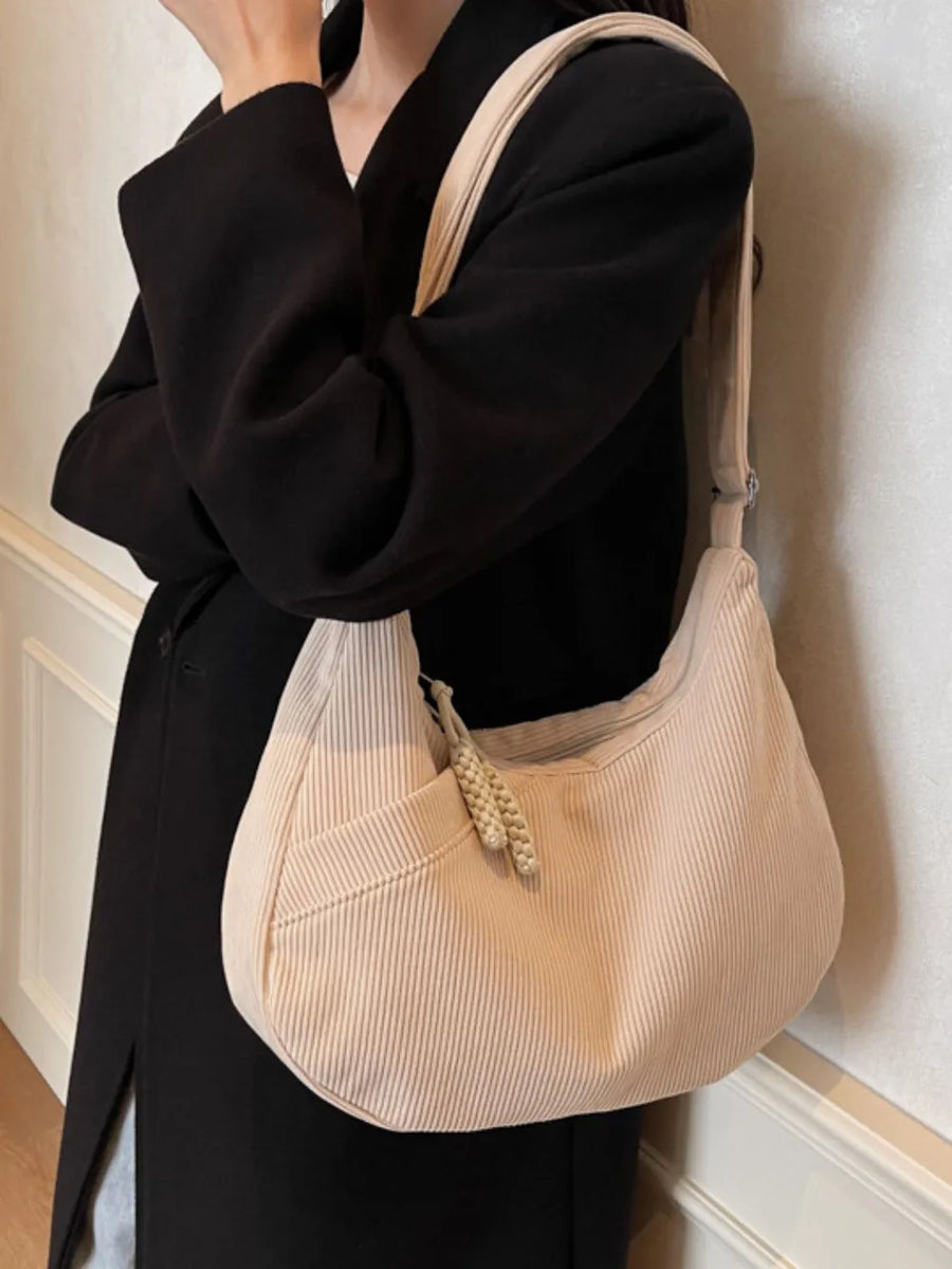 Beige corduroy crescent shoulder bag with braided tassel detail and front pocket.