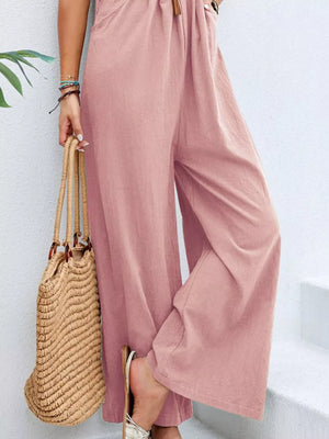 Pink wide-leg romper with pockets and adjustable drawstring waist, styled with sandals and a straw bag.