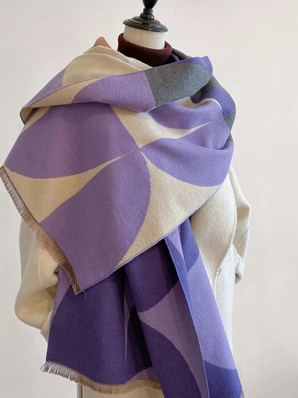 Soft geometric patterned scarf in purple and cream with fringes, styled on a mannequin.