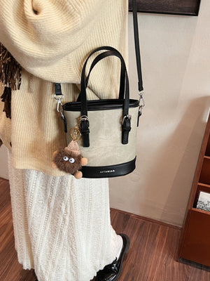 Two-tone bucket crossbody bag in beige and black with fuzzy charm and top handles.