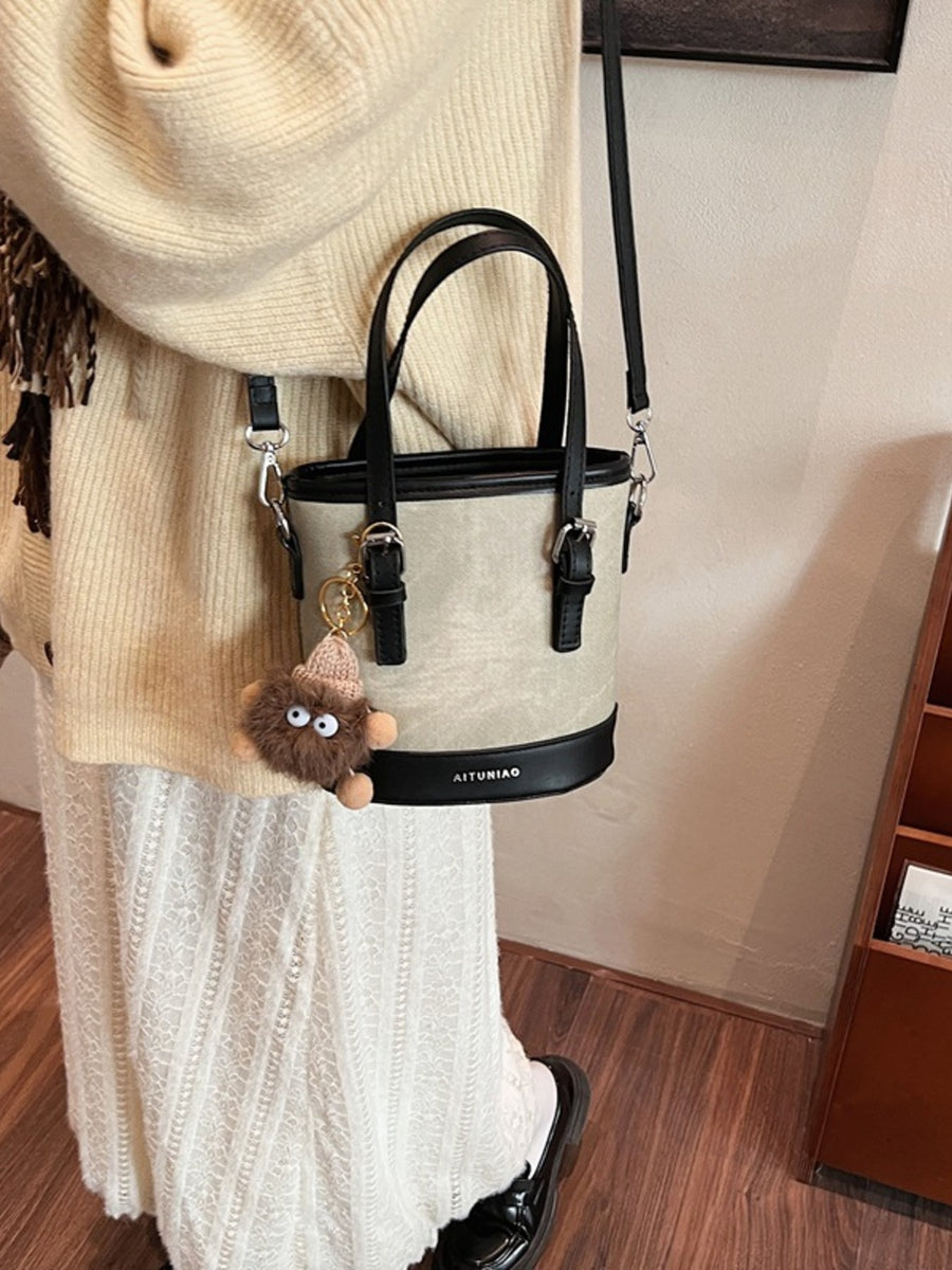 Two-tone bucket crossbody bag in beige and black with fuzzy charm and top handles.