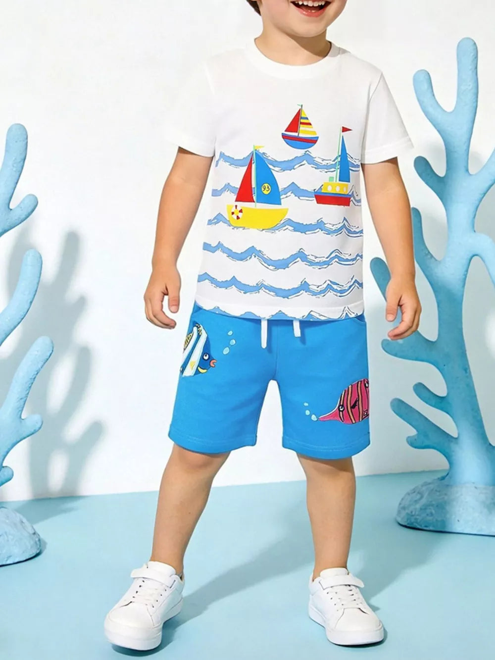 White cotton tee with sailboat print and blue shorts with fish print for boys.
