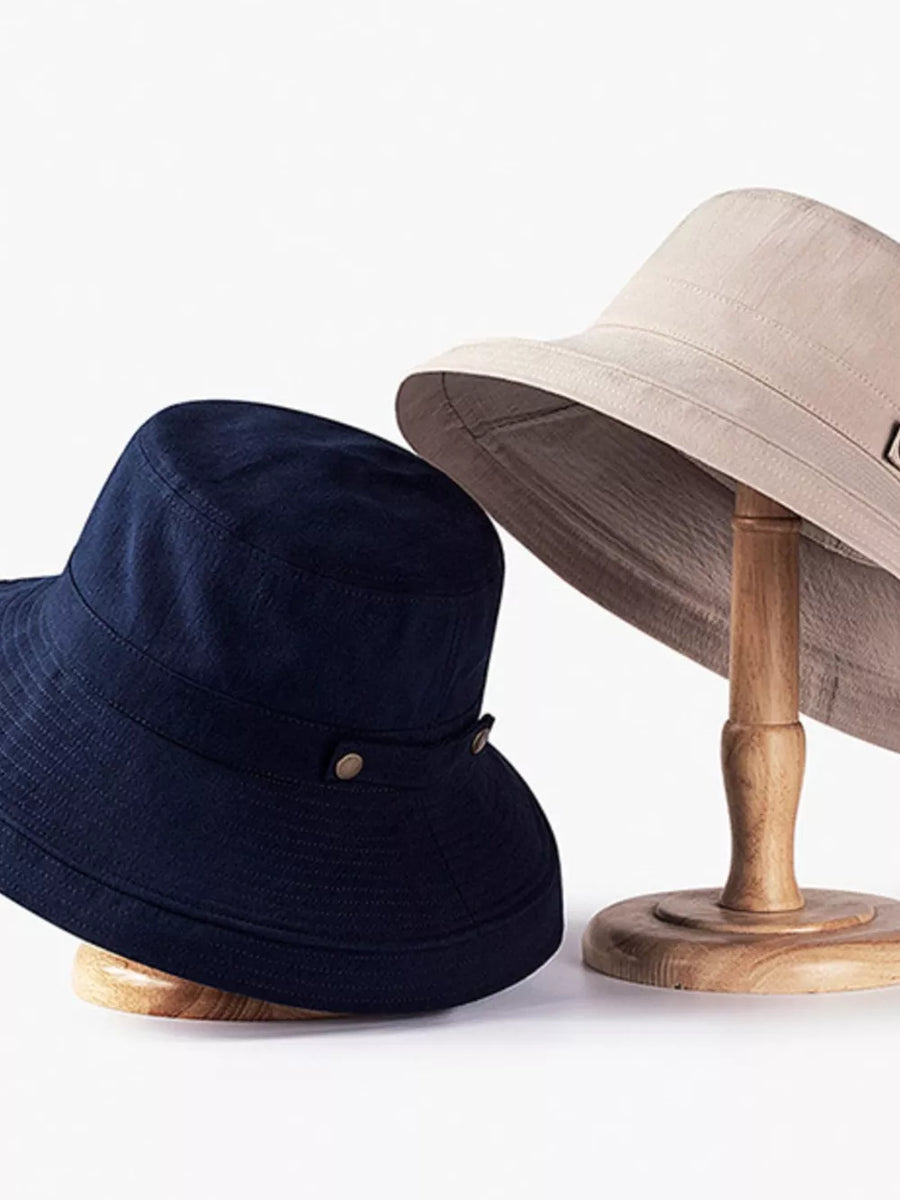 Dark navy cotton bucket hat with brass hardware detail next to a khaki wide brim sun hat.