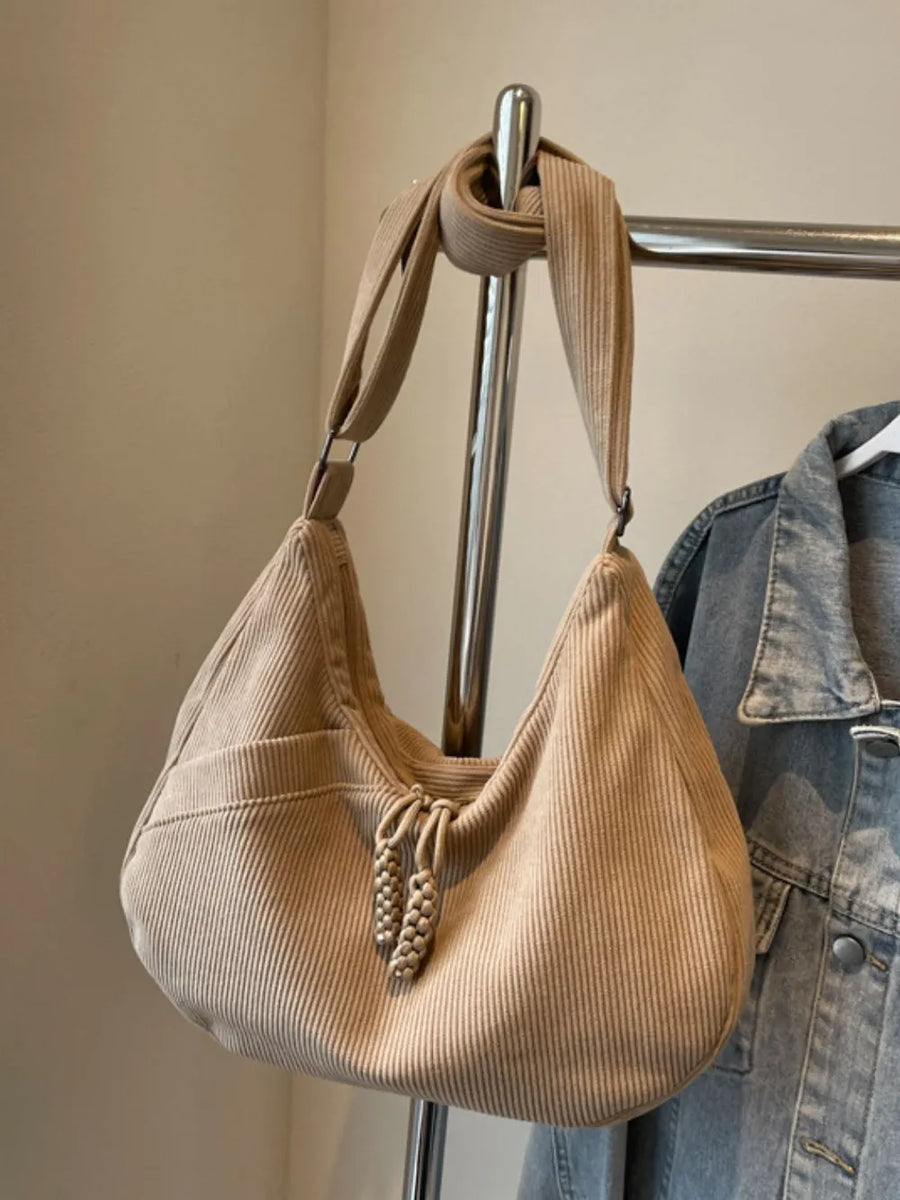 Beige corduroy crescent shoulder bag with braided tassels and front pocket.