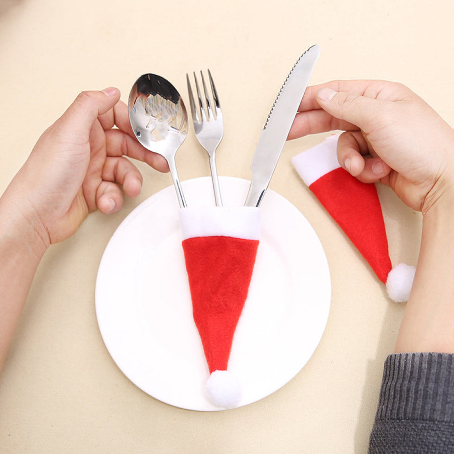 Red Santa hat cutlery holders with white trim and pom-poms, holding silverware on a plate.
