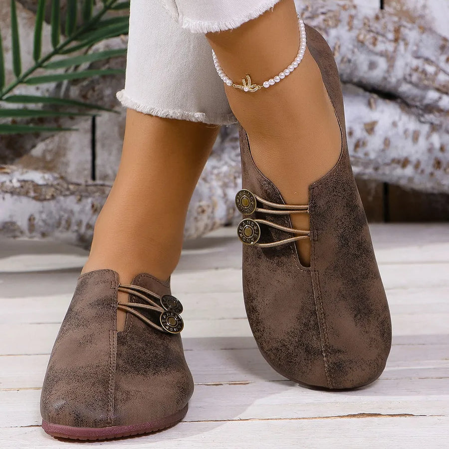 Brown leather slip-on flats with antique gold button details and elastic straps.