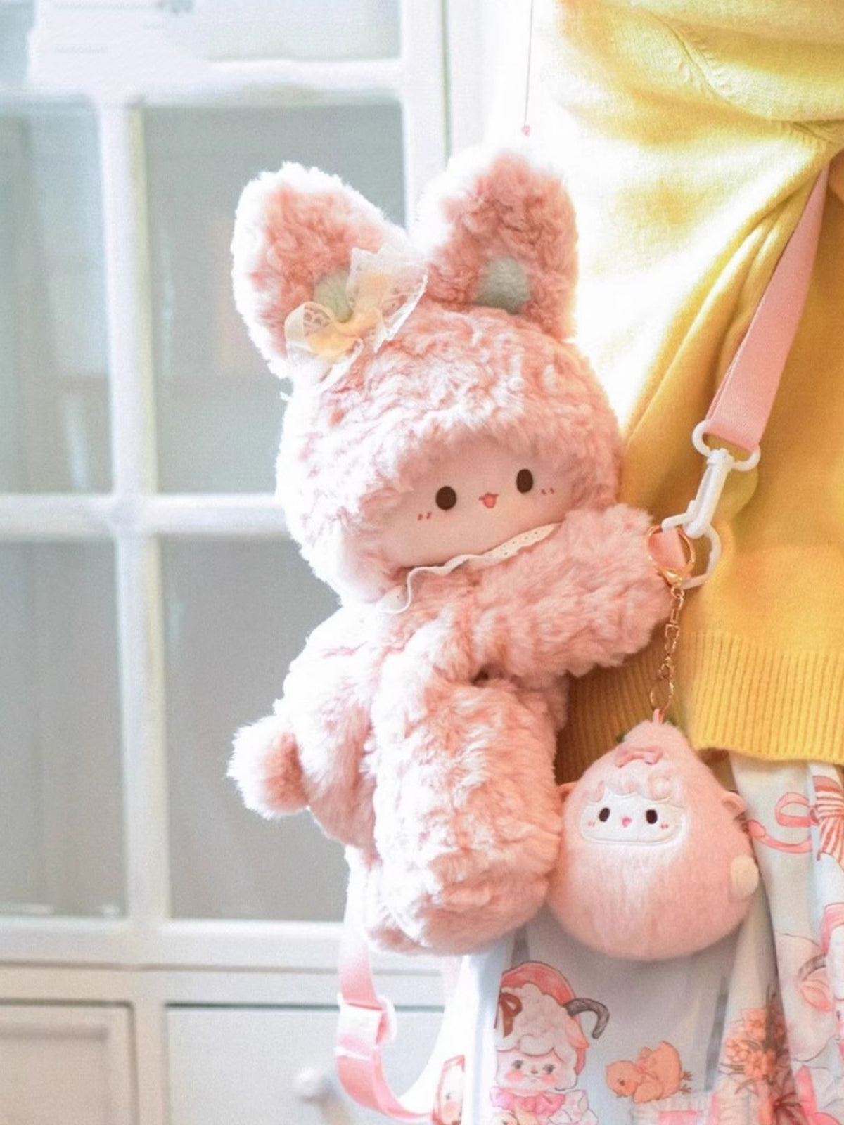 Pink plush bunny backpack with lace trim and a small matching pouch, worn by a person in a yellow sweater.