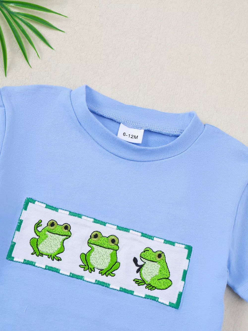 Light blue cotton baby t-shirt with three embroidered frogs on a white patch with green trim.