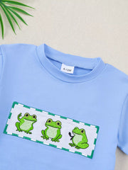 Light blue cotton baby t-shirt with three embroidered frogs on a white patch with green trim.