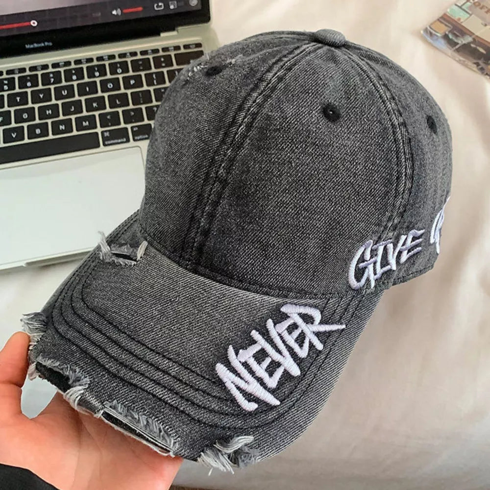 Distressed denim baseball cap in black with white embroidered text "NEVER GIVE UP" and frayed details.