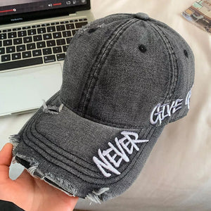 Distressed denim baseball cap in black with white embroidered text "NEVER GIVE UP" and frayed details.
