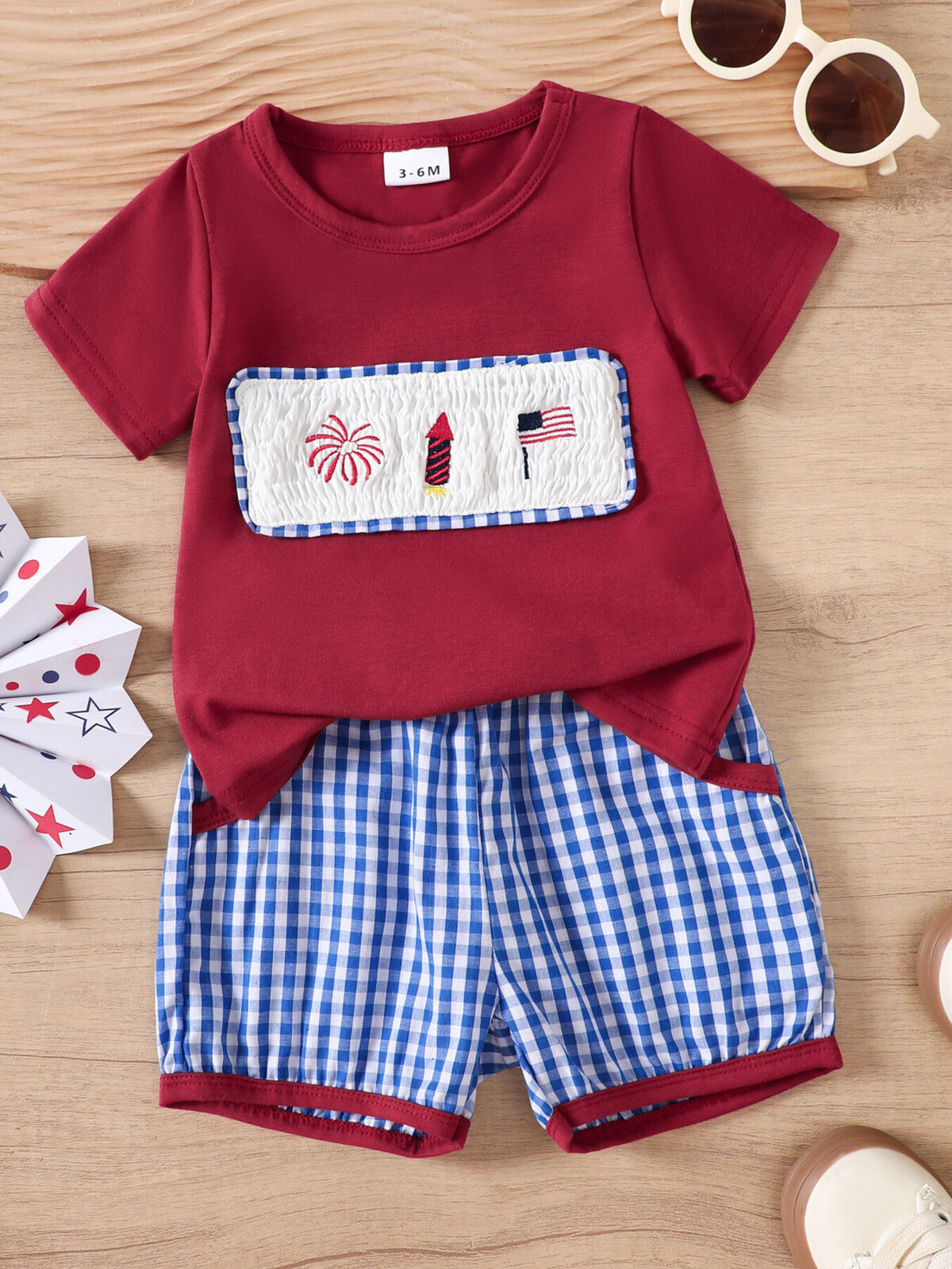 Maroon baby set with smocked panel featuring fireworks, rocket, and American flag embroidery, paired with blue gingham