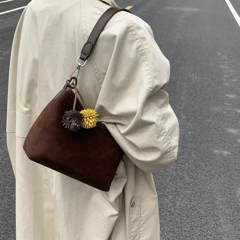 Brown suede crossbody bag with two pompom charms and a dark brown strap.